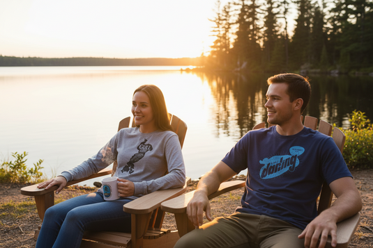 Two people sitting in Adirondack chairs by a lake at sunset, wearing Lakes Supply Co t-shirts and BWCA coffee mug.